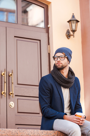 Enjoying hot drink. Handsome young man in smart casual wear sitting in front of the house entrance and holding coffee cupの写真素材