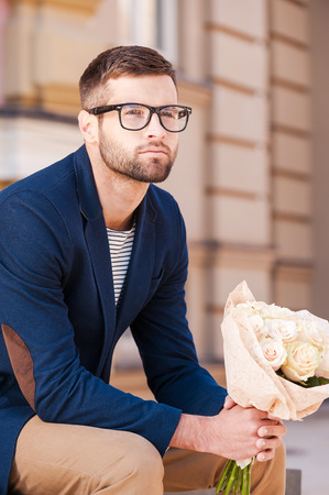 The freshest roses for her. Handsome young man in smart jacket holding bouquet of flowers and looking away while sitting on the bench outdoorsの写真素材