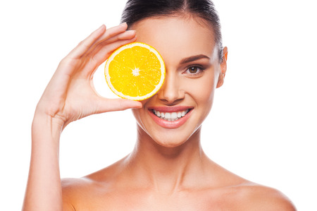 Great food for a healthy lifestyle. Beautiful young shirtless woman holding piece of orange in front of her eye while standing against white の写真素材