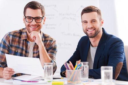 Creative colleagues. Two cheerful businessmen in smart casual wear sitting together at the table and looking at cameraの写真素材