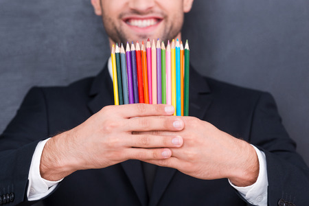 He got creative mind.  Close-up of colorful pencils in the hands of smiling businessman standing against blackboardの写真素材
