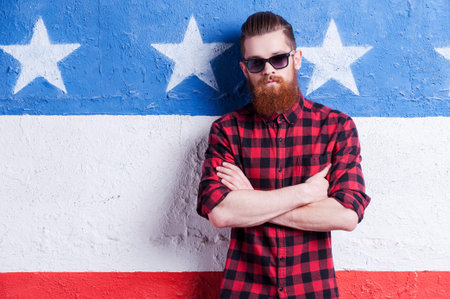 American style. Handsome young bearded man in sunglasses keeping arms crossed and looking at camera...