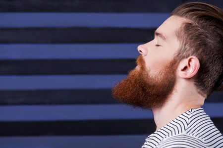 Perfect beard. Side view of handsome young bearded man holding hand on chin while standing against stripped backgroundの写真素材