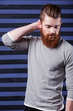 Casual and cool. Side view of handsome young bearded man holding hand on head while standing against stripped backgroundの写真素材