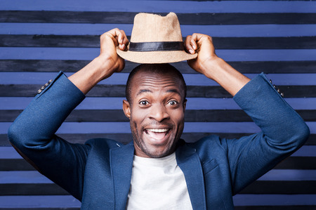 Funky style. Handsome young African man holding his hat upon his head and smiling while standing against striped backgroundの写真素材
