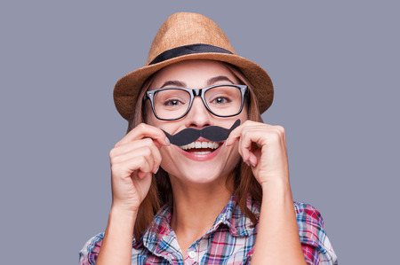 Having fun with face mustache. Cheerful young woman in funky hat holding fake mustache on her face and looking at camera while standing against grey backgroundの写真素材