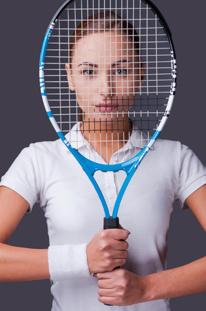 I know how to win! Beautiful young women in sports clothes holding tennis racket in front of her face and looking at camera while standing against grey backgroundの写真素材