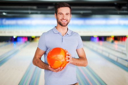 Bowling master. Cheerful young man holding a bowling ball and smiling at camera while standing against bowling alleysの写真素材