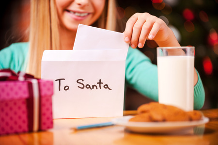 Letter to Santa. Close-up of cheerful little girl putting a letter to Santa into the envelope while sitting at the table with glass of milk and cookies laying on itの写真素材
