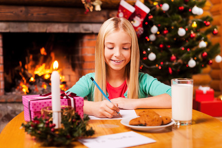 Writing a letter to Santa. Cute little girl writing a letter to Santa Claus while sitting at home with Christmas tree and fireplace in the backgroundの写真素材