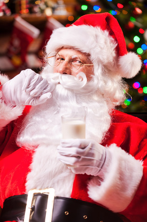I love milk! Cheerful Santa Claus holding glass with milk and adjusting his mustache with Christmas Tree in the backgroundの写真素材