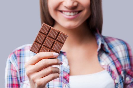 Chocolate mood. Close-up of young women holding bar of chocolate and smiling while standing against grey backgroundの写真素材