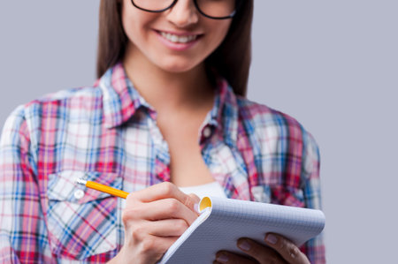 Making notes. Close-up of young women writing in notebook by a pencil while standing against grey...