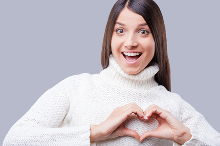 Beauty in love. Happy young women in warm winter clothing keeping hands in a shape of heart while standing against grey backgroundの写真素材