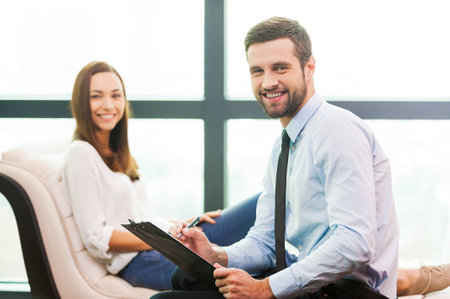 Always ready to help you. Confident young man in shirt and tie holding clipboard and smiling while woman sitting in the backgroundの写真素材