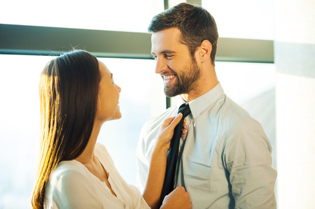 Now you are ready! Beautiful young loving couple standing face to face and smiling while woman adjusting necktie of her husbandの写真素材