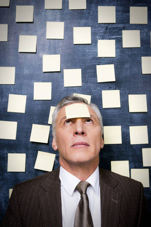 Frustrated senior man in formalwear with adhesive note on his forehead looking up while standing against blackboardの写真素材