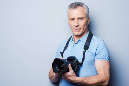 Portrait of senior mature man in T-shirt holding camera while standing against grey backgroundの写真素材