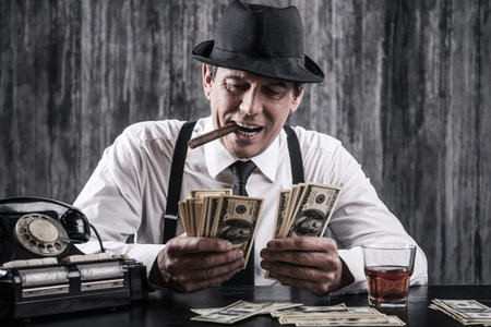 Money and power. Senior gangster in shirt and suspenders counting money and smiling while sitting at...