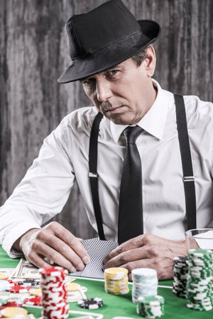 So what you got for me? Serious senior man in shirt and suspenders sitting at the poker table and holding cards  with money and gambling chips laying all around himの写真素材