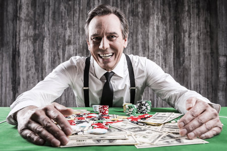 It is all mine! Happy senior man in shirt and suspenders sitting at the poker table and embracing chips and money laying on itの写真素材
