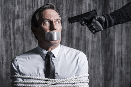 Please no. Tied up businessman with adhesive tape on his mouth sitting in front of the dirty wall while someone aiming his headの写真素材