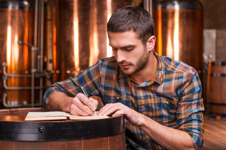 Planning his beer business. Confident young man in casual shirt writing something in his note pad while leaning at the wooden barrel with metal containers in the backgroundの写真素材