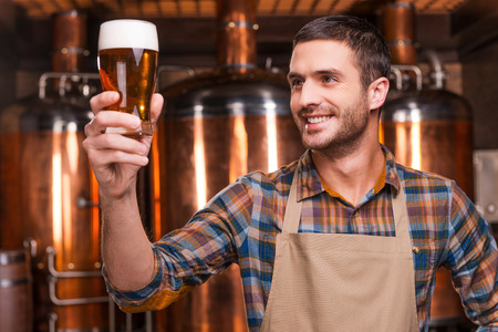 Happy brewer. Happy young male brewer in apron holding glass with beer and looking at it with smile while standing in front of metal containersの写真素材
