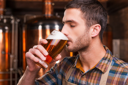 Tasting fresh brewed beer. Handsome young male brewer in apron tasting fresh beer and keeping eyes closed while standing in front of metal containers