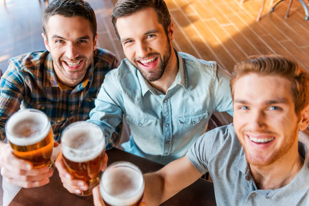 Cheers! Top view of three happy young men in casual wear toasting with beer while sitting in bar togetherの写真素材
