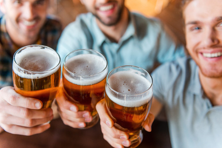 Cheers to success! Top view of three happy young men in casual wear toasting with beer while sitting in bar together