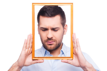  Portrait of frustrated young man in shirt holding picture frame in front of his face  while standing against white backgroundの写真素材