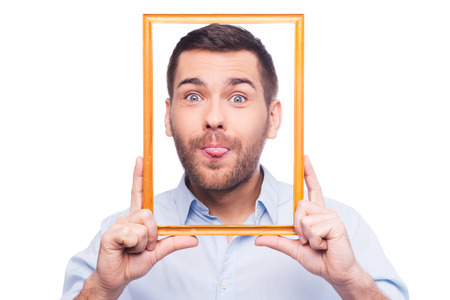 Playful portrait Handsome young man in shirt sticking his tongue out and holding picture frame in front of his face while standing against white backgroundの写真素材