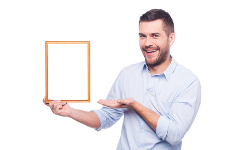 Handsome young man in shirt holding a picture frame and pointing it with smile while standing...