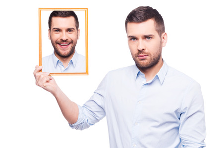Handsome young man in shirt holding a picture of himself with smiling face and looking sad while standing against white backgroundの写真素材
