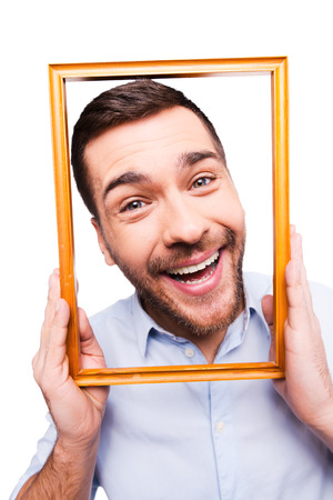 Handsome young man in shirt holding picture frame in front of his face and smiling while standing against white backgroundの写真素材