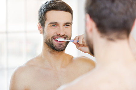 Brushing teeth in the morning. Rear view of handsome young beard man brushing his teeth and smiling while standing against a mirrorの写真素材