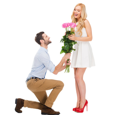 They are as beautiful as you! Full length of man standing at his knee and giving a bouquet of pink roses while both isolated on white backgroundの写真素材