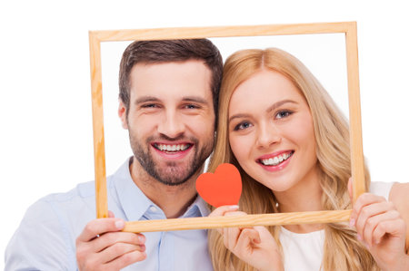 Celebrating their love. Beautiful young loving couple bonding to each other and holding heart shape paper while looking through a frame and standing isolated on white backgroundの写真素材