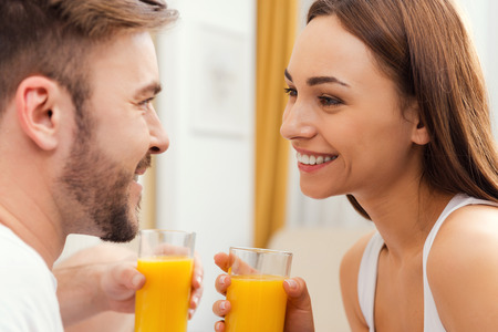 Enjoying fresh juice together. Beautiful young loving couple holding glasses with orange juice and looking at each other with smileの写真素材