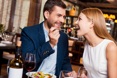 Try this! Beautiful young loving couple enjoying dinner at the restaurant while man feeding his girlfriend with saladの写真素材