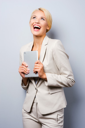 Young and successful businesswoman. Beautiful young businesswoman holding digital tablet and looking up while standing against grey backgroundの写真素材