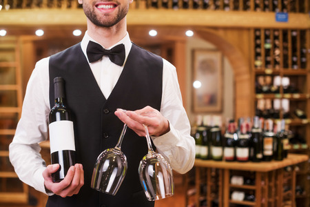 Confident waiter. Handsome young man in waistcoat and bow tie holding bottle and glasses while standing in liquor storeの写真素材