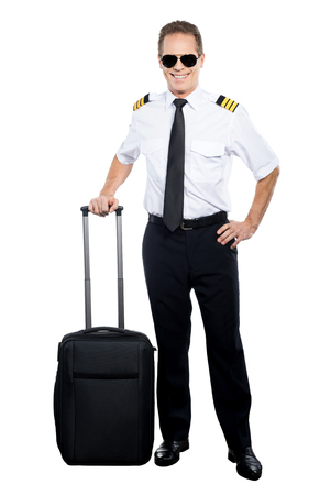 New day and new destination. Confident male pilot in uniform leaning hand on his suitcase and smiling while being isolated on white backgroundの写真素材