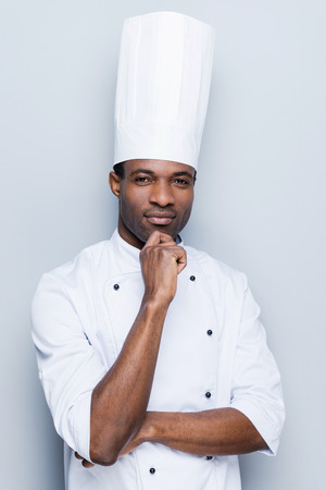 Confident and experienced chef. Confident young African chef in white uniform holding hand on chin and looking at camera while standing against grey backgroundの写真素材