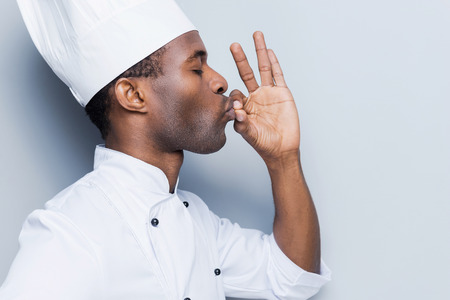 This meal is just perfect! Side view of confident young African chef in white uniform keeping eyes closed and gesturing while standing against grey backgroundの写真素材