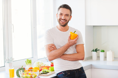 Healthy food is healthy life. Handsome young man holding fresh yellow pepper and smiling while standing in the kitchenの写真素材
