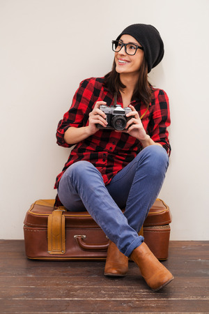 Full of inspiration.  Beautiful young woman in headwear holding camera and looking away while sitting on suitcaseの写真素材