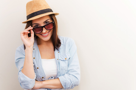 Flirting with you. Portrait of beautiful young woman in glasses and funky hat adjusting her glasses and smiling while standing against brown backgroundの写真素材