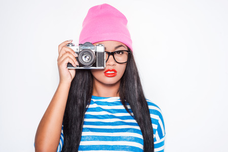 Focusing at you. Attractive young African woman in funky clothes focusing at you with her old-fashioned camera while standing against white backgroundの写真素材